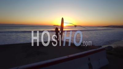 Aerial View Over A Woman Standing With A Surf Board Looking Out At The Sunset - Video Drone Footage
