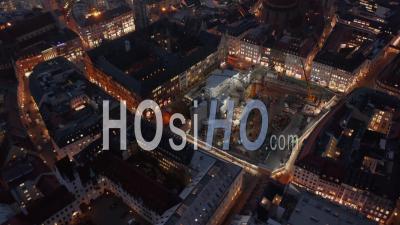 Huge Construction Building Site In Urban Environment City Center Of Munich, Germany At Night With Lighting Equipment And Big Machinery, Aerial Dolly Out Tilt Up - Video Drone Footage