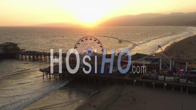 Close Up View Of Santa Monica Pier Ferris Wheel, Los Angeles At Beautiful Sunset With Tourists, Pedestrians Walking Having Fun At Theme Park Rollercoaster With Ocean View Waves Crashing 4k - Video Drone Footage